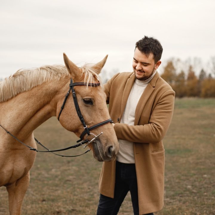 Brunette man with beard and brown horse standing in the field. Man wearing beige coat. Man touching the horse.